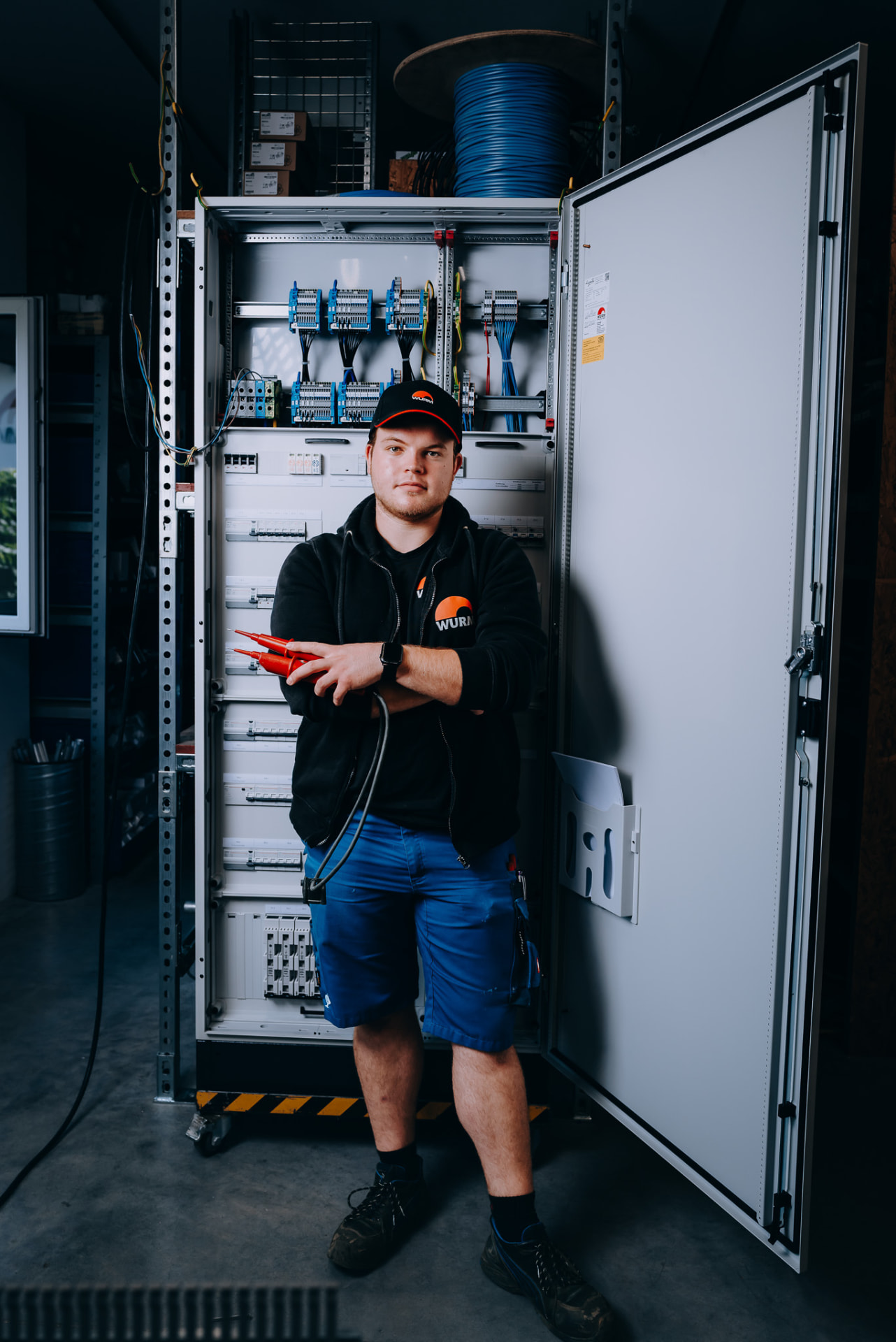 Elektroniker mit Prüfspitzen vor geöffnetem Schaltschrank